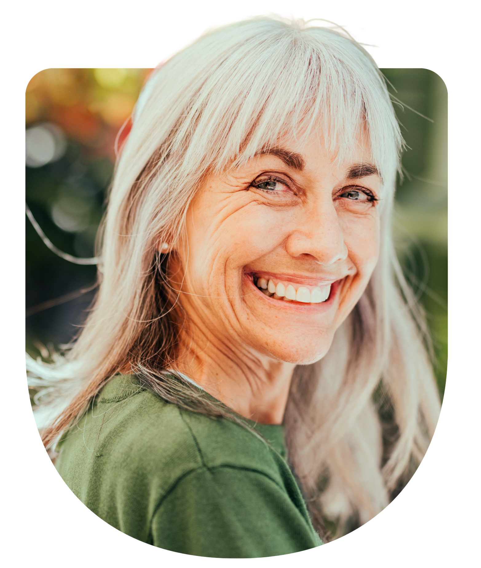 Older woman white white hair and green shirt smiling in the sun with blurry nature behind her